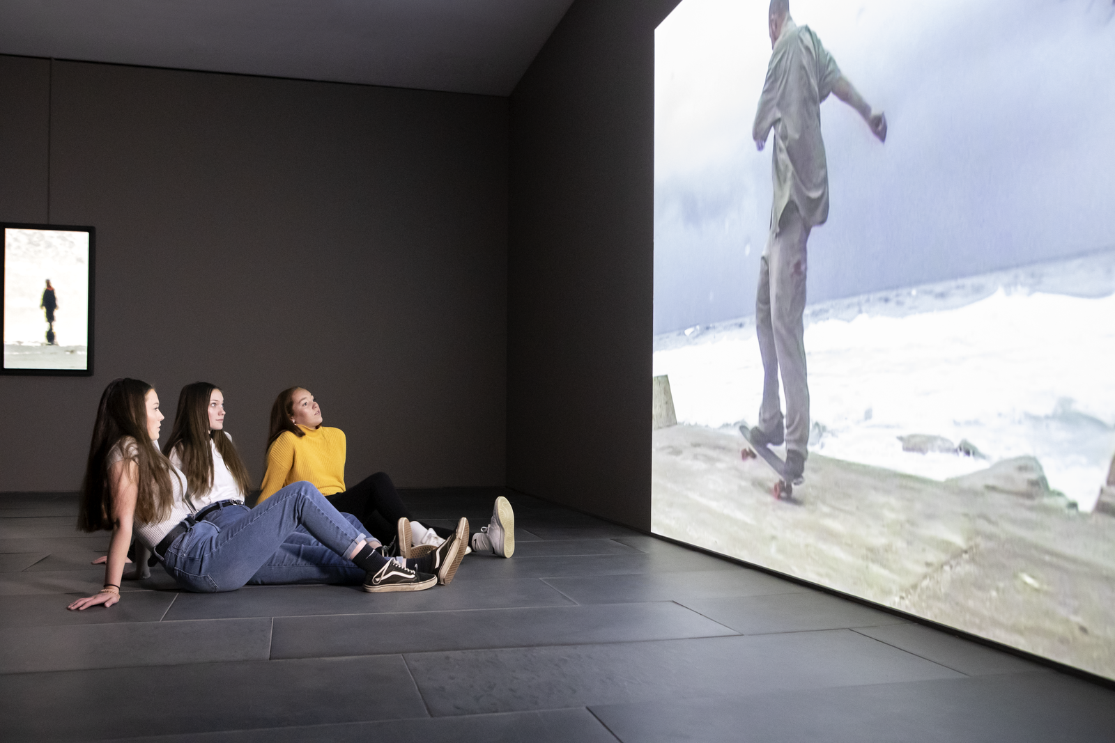 Three teenagers sit on the floor and watch a large audio visual installation projected onto a wall