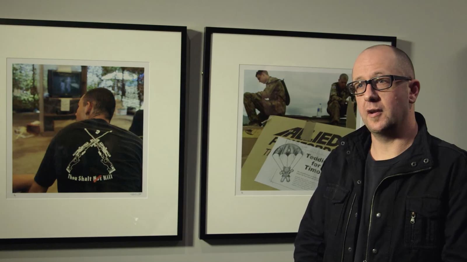 A video still of a man speaking to the camera in front of two artworks