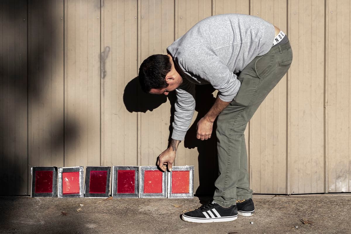 James Tylor, in olive chinos, grey knit and black and white Adidas trainers, reaches down to touch one of 6 daguerreotypes exposing in the sunshine
