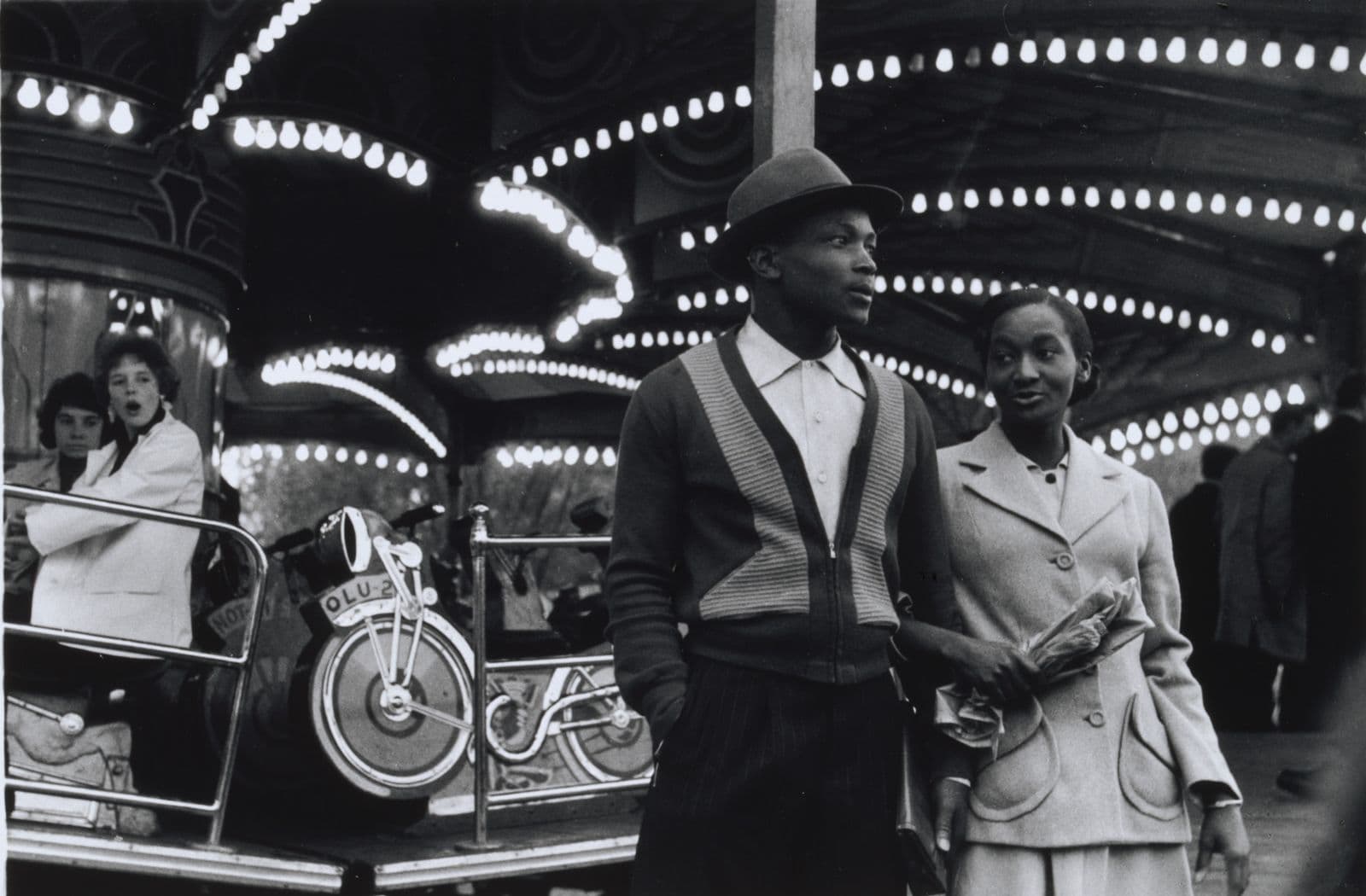 Black and white photograph of a man and a woman with linked arms in front of a carousel. In the background two women look on, one with a shocked expression