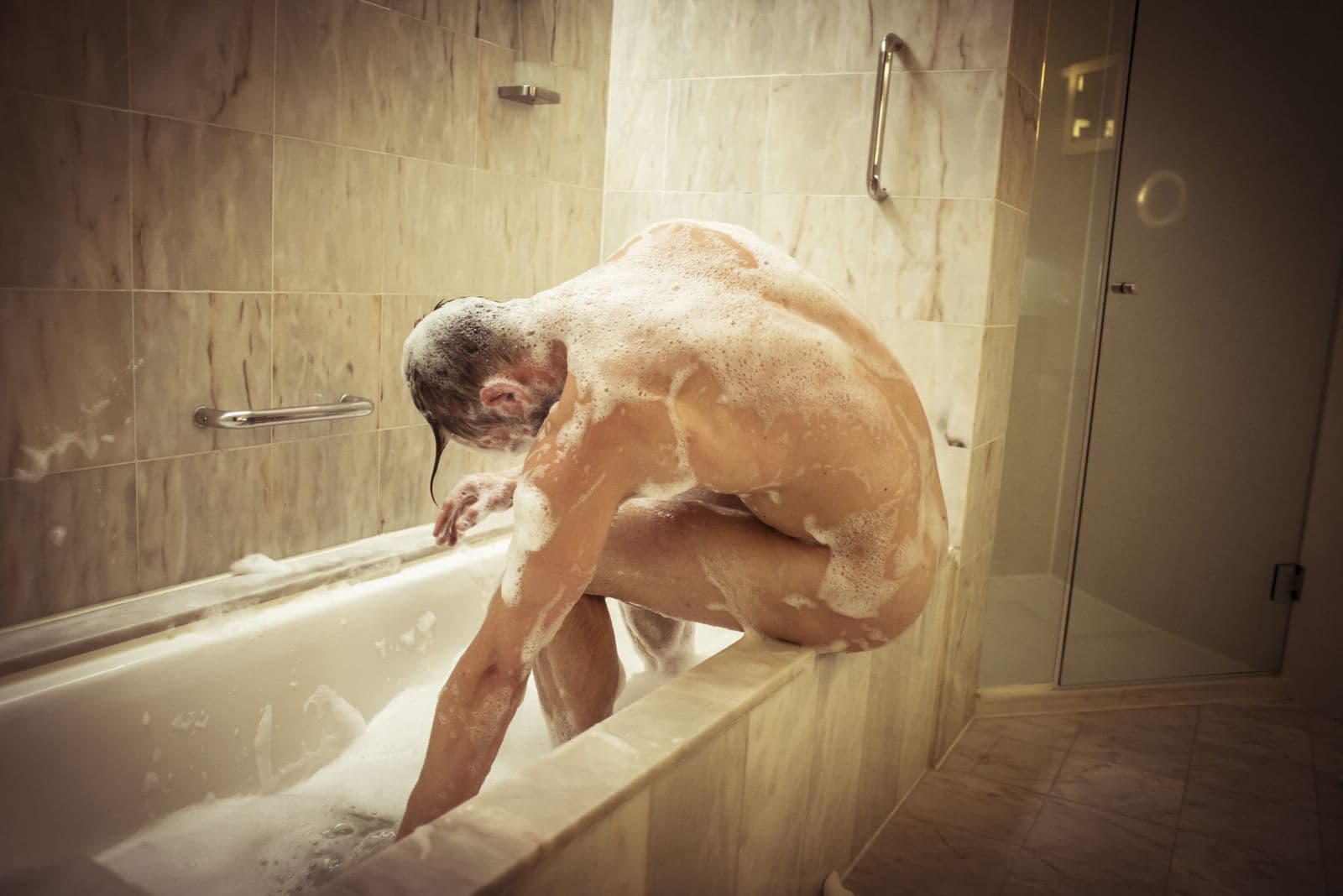 Photo of a man sitting on the side of a bathtub covered with soapy foam