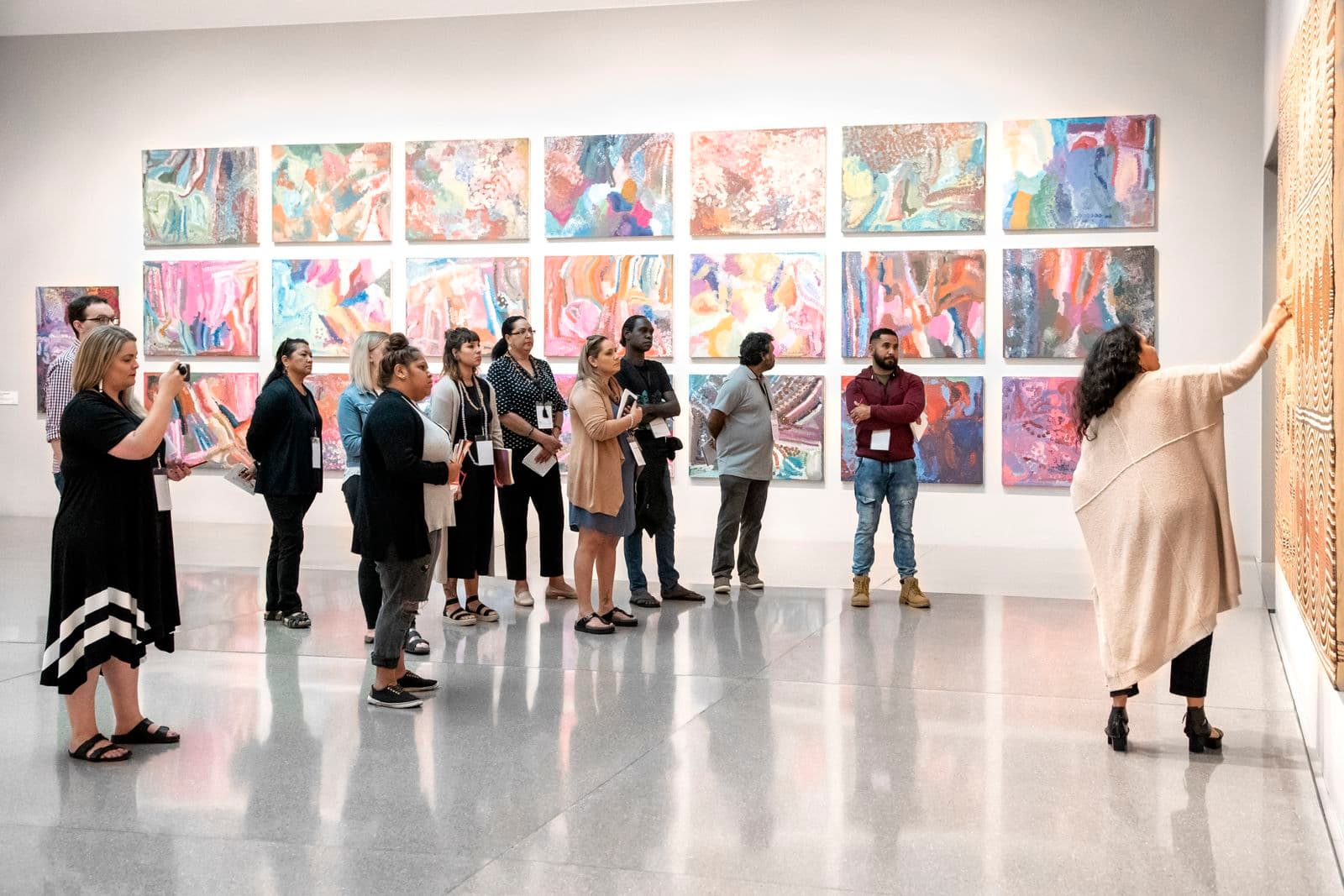 A group of people are standing in an arc around a painting in a gallery space with one person speaking about at pointing at the painting