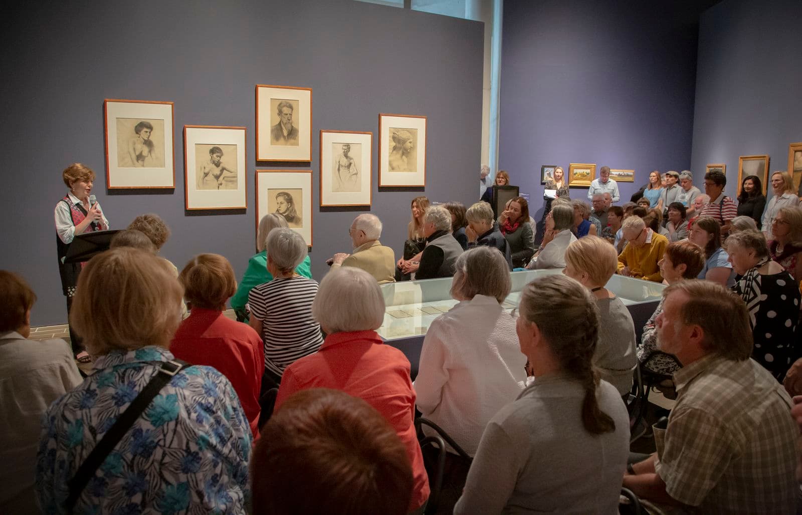 In a gallery space a woman is standing at a lectern speaking to a room of seated people.