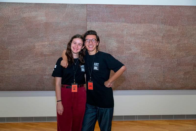 A teenage girl and boy wearing glasses with his arm around her shoulder. Both smiling and standing in front of artwork.