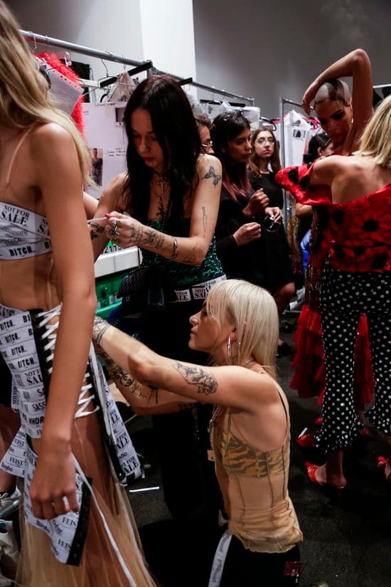 In a crowded room behind the scenes at a fashion runway show, two women are standing and kneeling behind a model wearing a dress, holding and pinning the fabric.