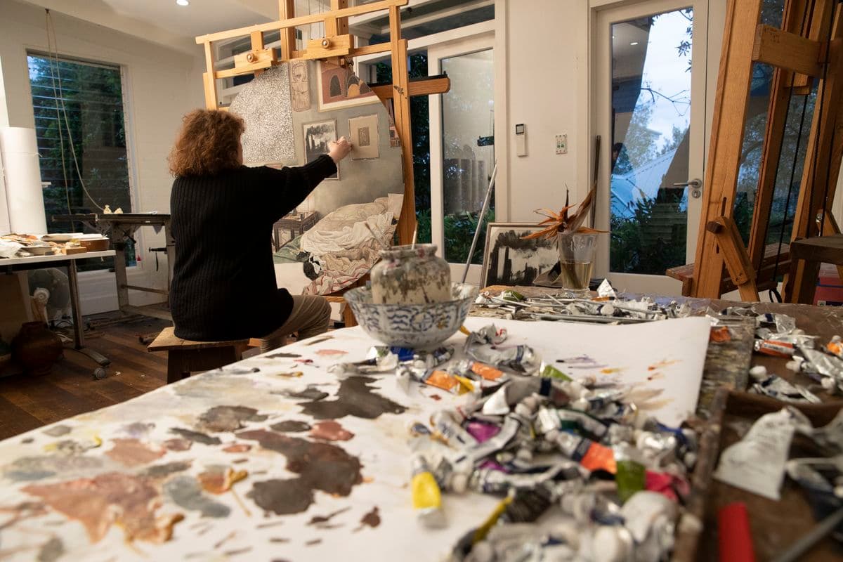 A view inside an art studio with a cluttered desk of paint tubes and a used palate. A woman artist is working on a work on a easel with her back towards the camera.