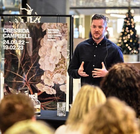 Photo of man giving a talk in front of Cressida Campbell exhibition poster