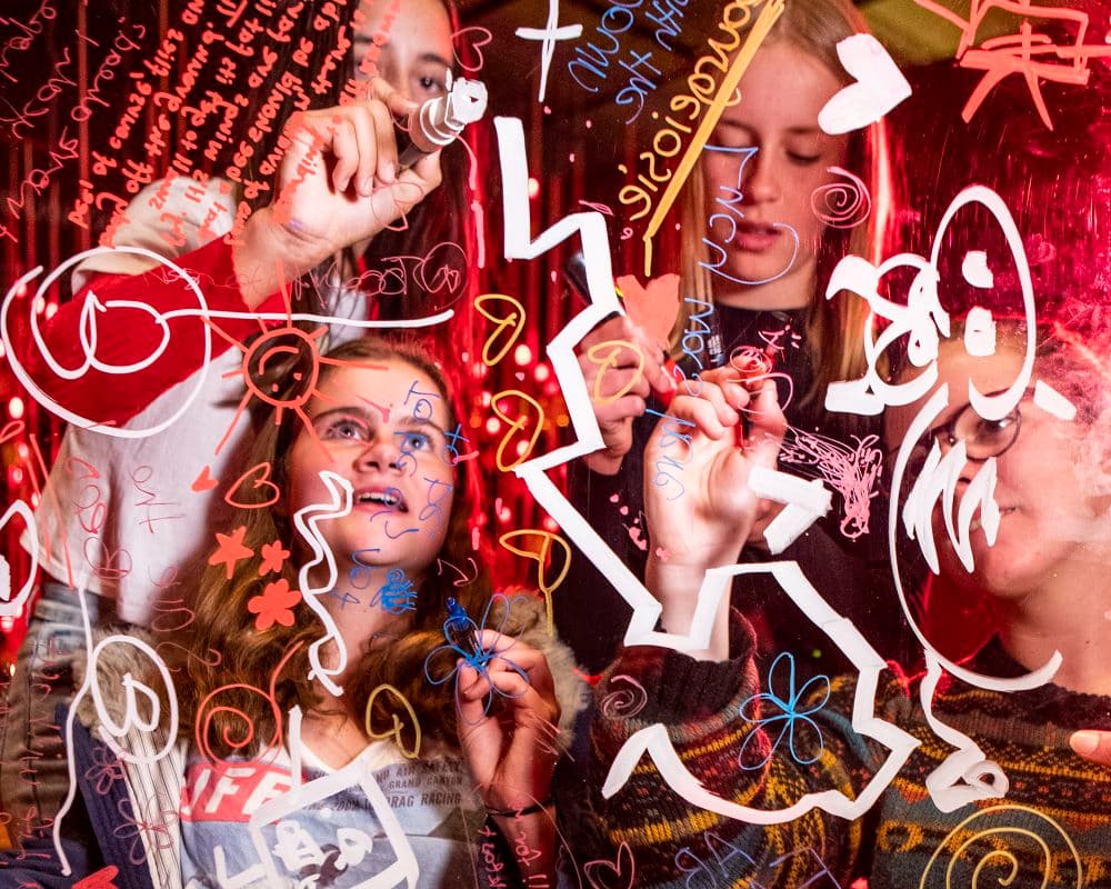 A group of people scribble with coloured markers on a clear screen