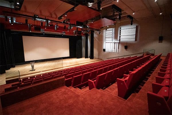 Photo of a Lecture theatre with fixed red chairs and a stage and large projection screen at the back