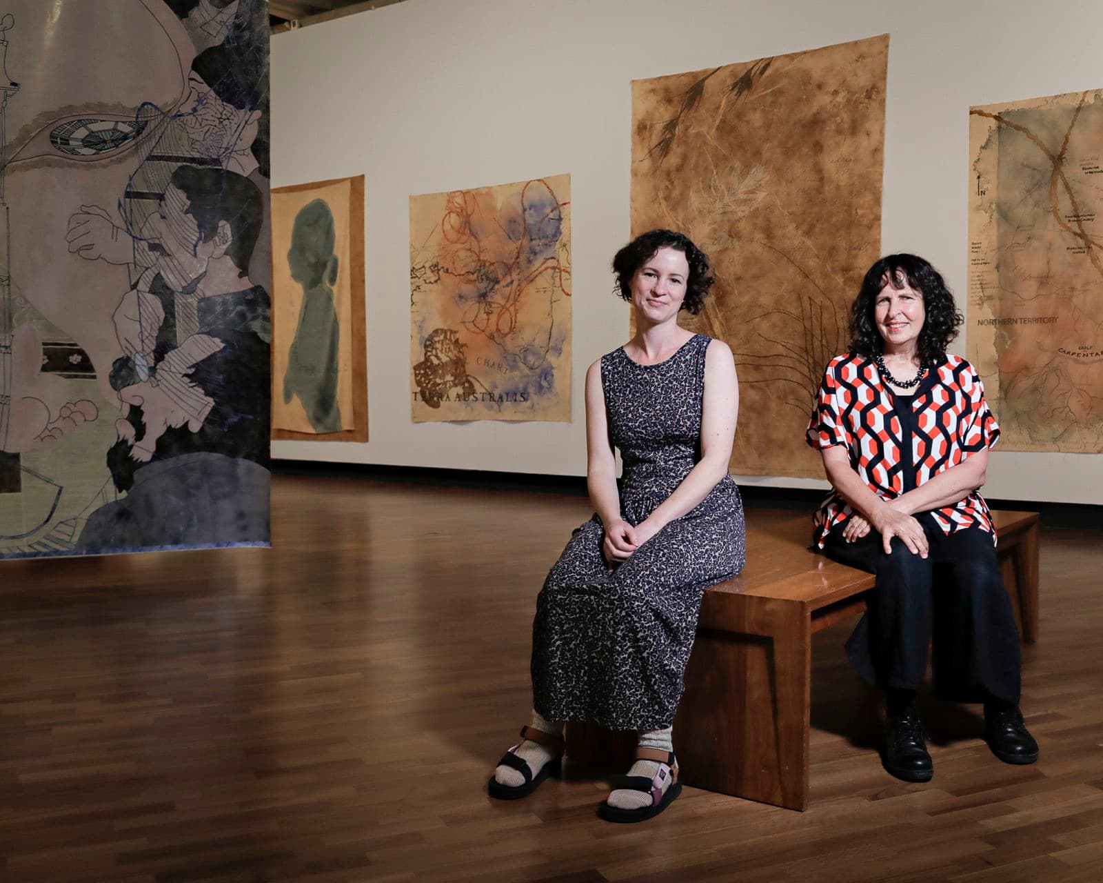 Two women sit on a timber bench, within a gallery space, surrounded by paintings and work of art suspended from the ceiling and walls