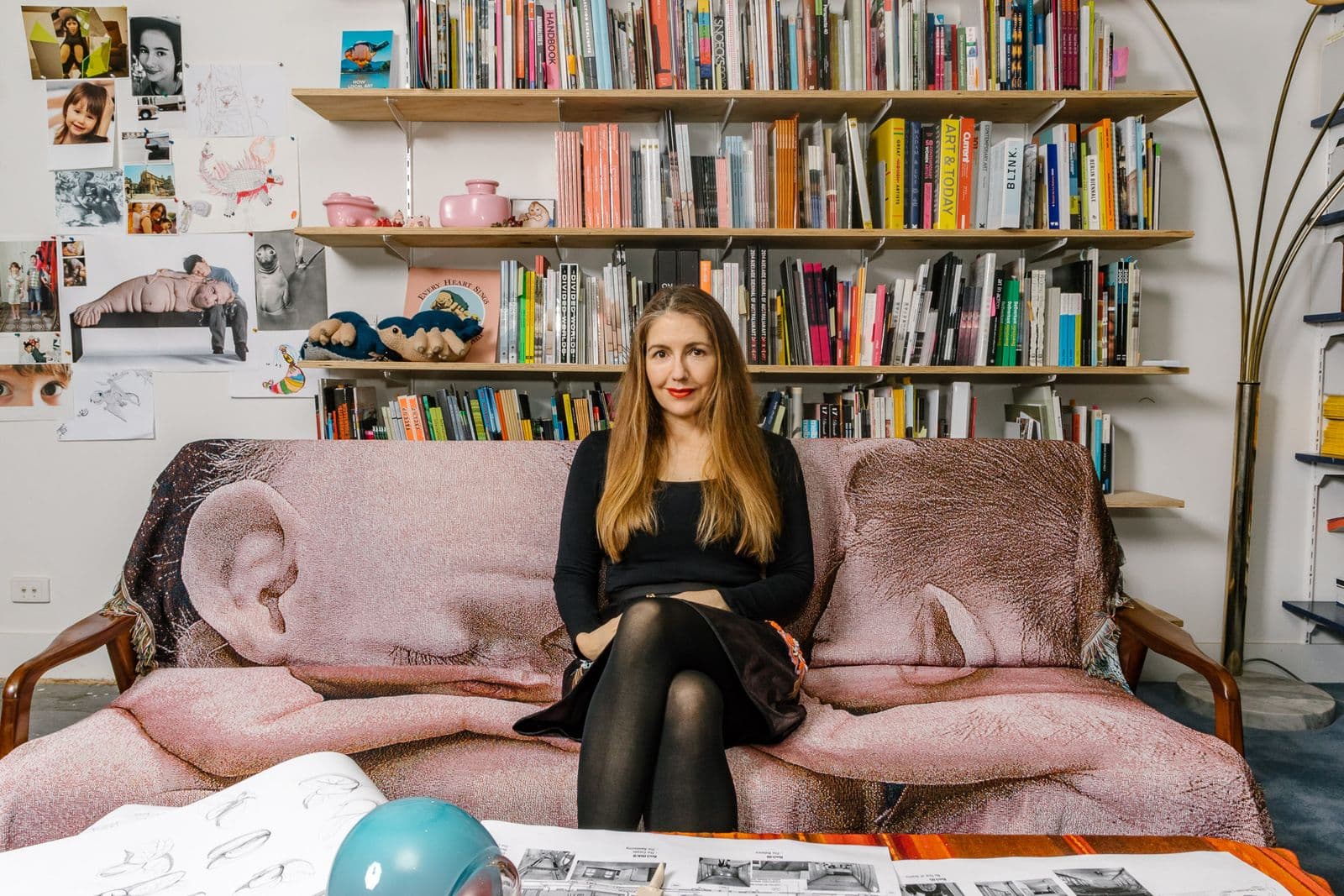 Artist Patricia Piccinini sits on a pink couch with a shelf of books in the background.