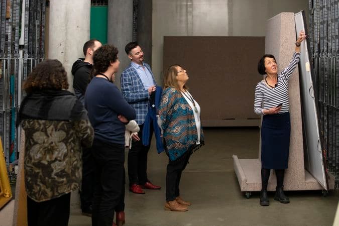 Five people are watching a demonstration given by a woman in a painting storage area.