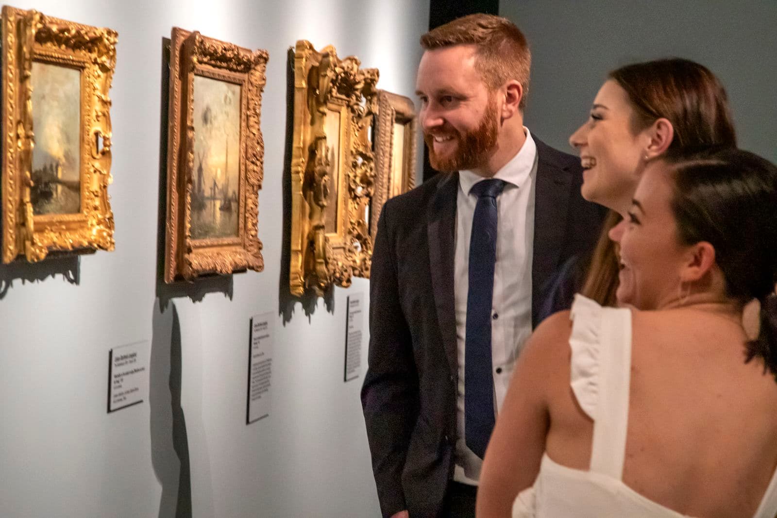 Two women and a man are smiling looking at works of art on a wall