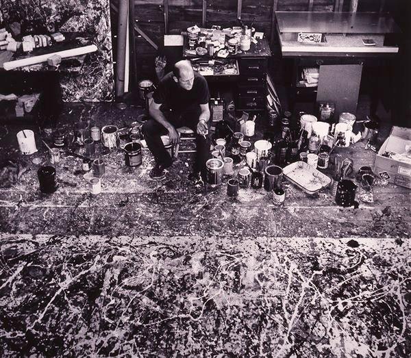 A white man sits in front of a messy abstract painting with paint tins to his side smoking a cigarette