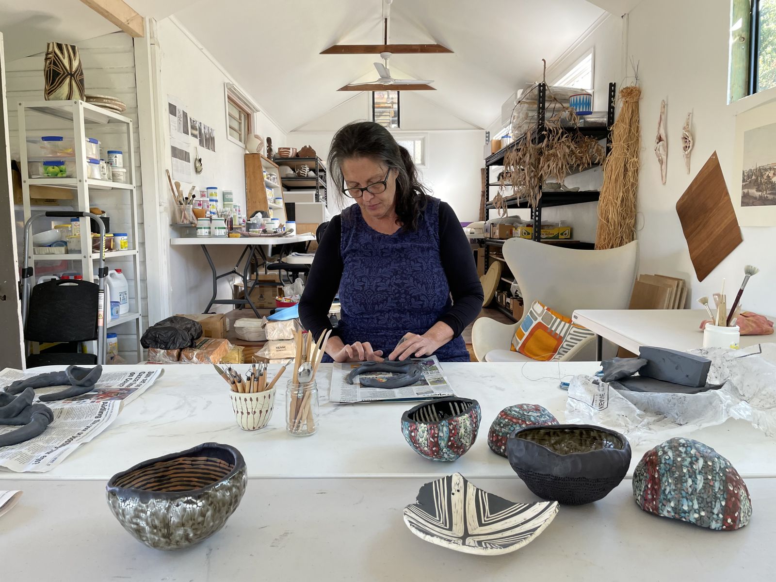 Penny Evans stands behind a large and full work bench making her artworks