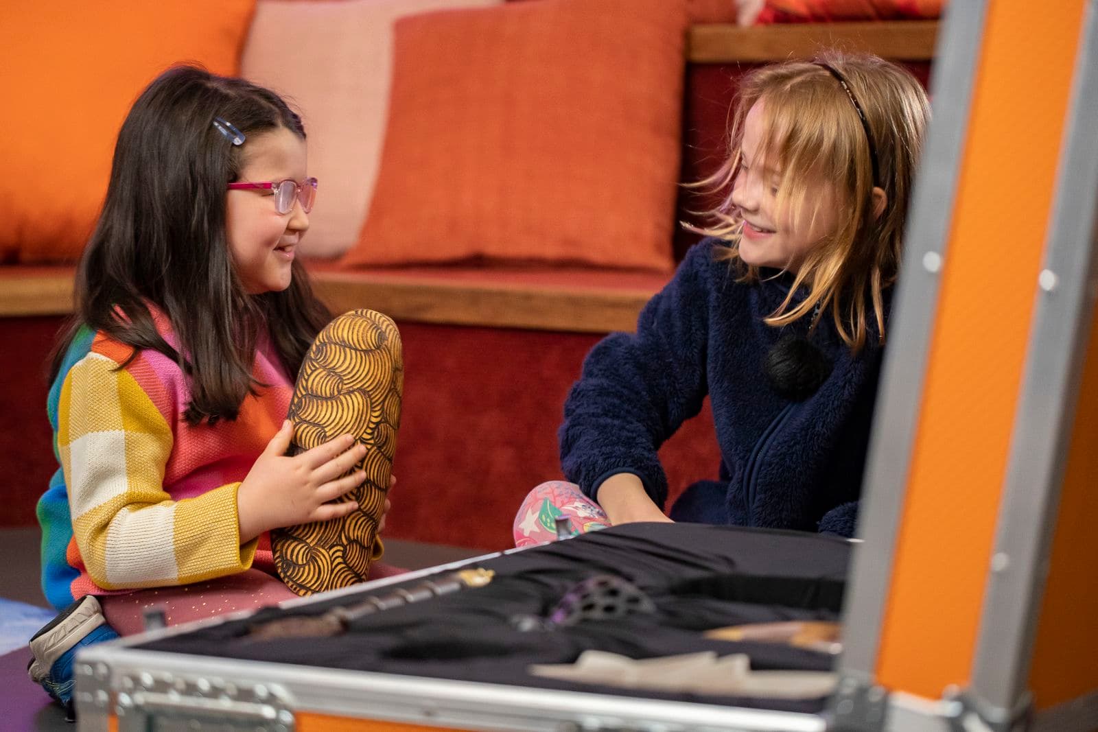 two young girls are smiling and holding objects they have taken out of a large orange case