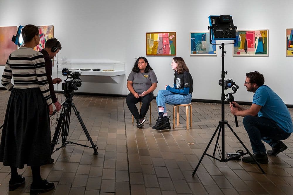 Two people sit on stools as three other people film them
