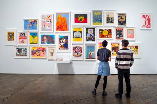 Two women stand looking at a wall hung with prints and paintings of various sizes.