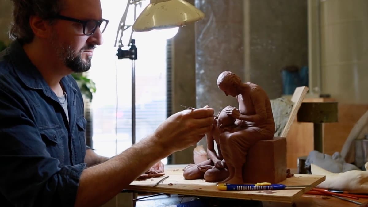 A video still of a man working on a small sculpture in the studio