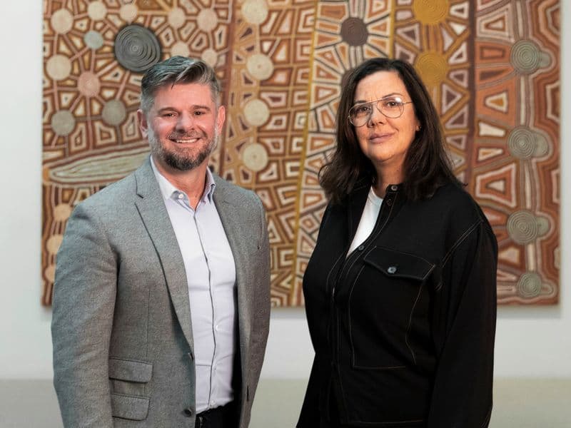 Hetti Perkins and Nick Mitzevich stand next to each other facing the camera with paintings from the First Nations gallery behind them