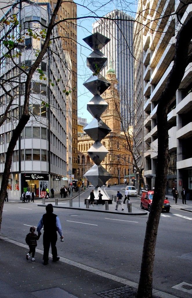 A large-scale sculptural work of seven steel trapezoidal prisms stacked vertically, and surrounded by buildings.