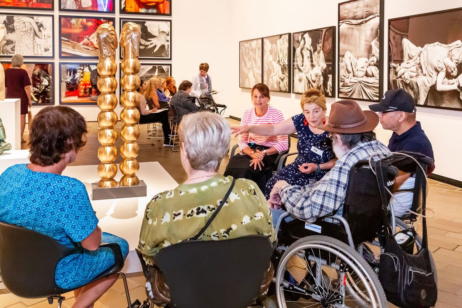 a group of people, some in wheelchairs, sit around a sculpture listening to a guide