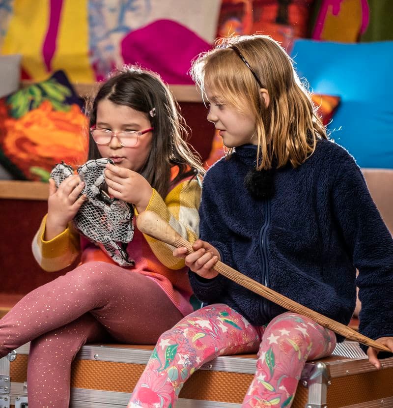 Two young girls are sitting on top of a large orange case, holding and examining objects
