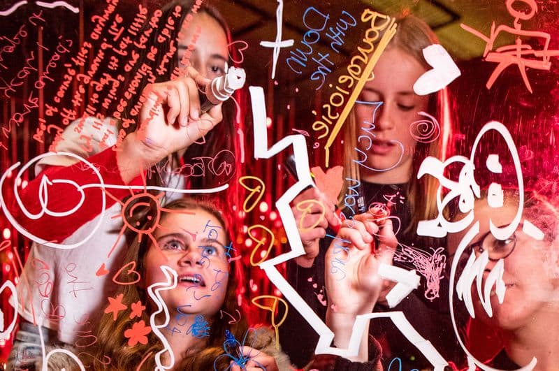 Young women are standing behind a piece of clear perspex, using markers to draw and write on it