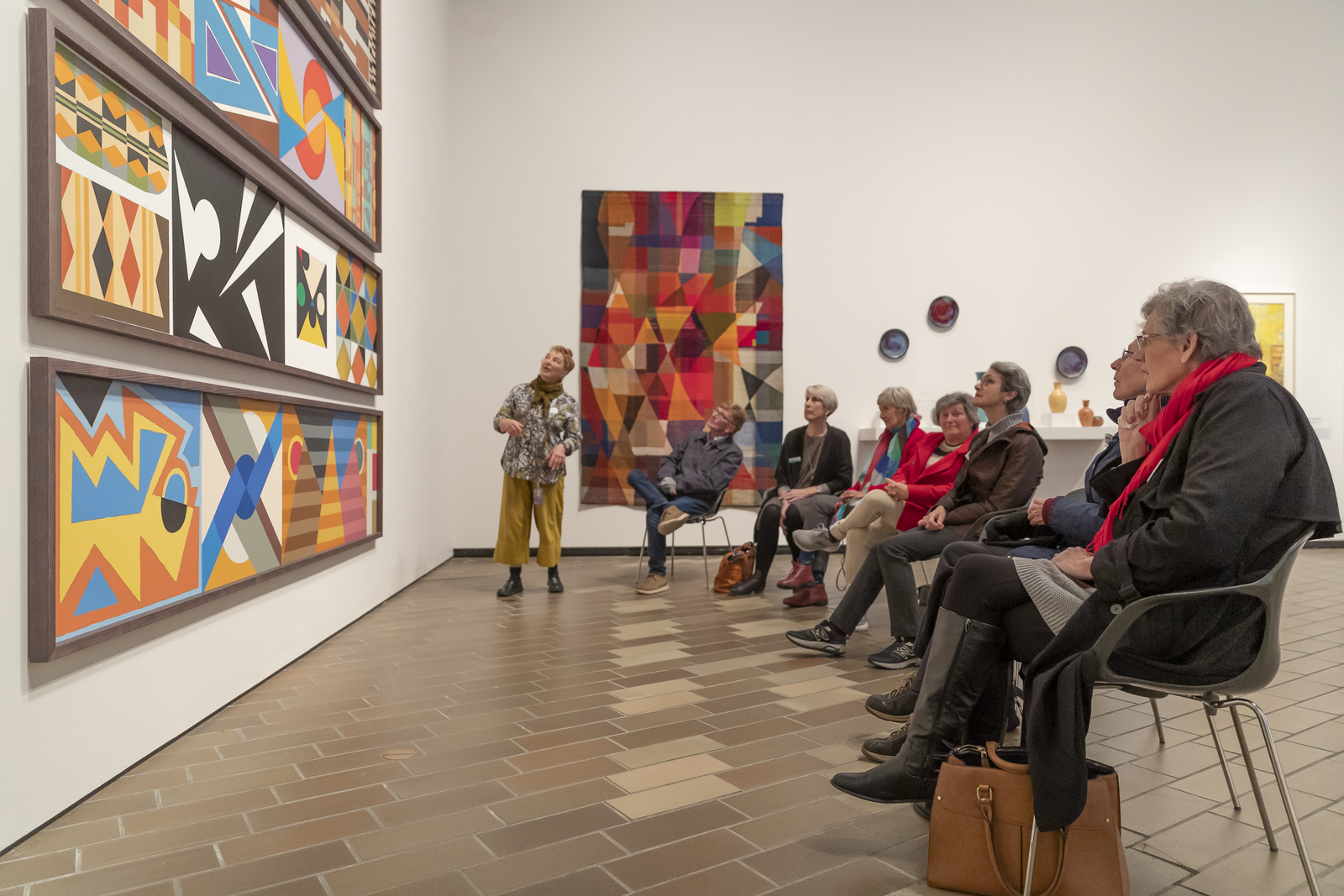 Group seated in front of colourful artwork with staff member standing and gesturing.