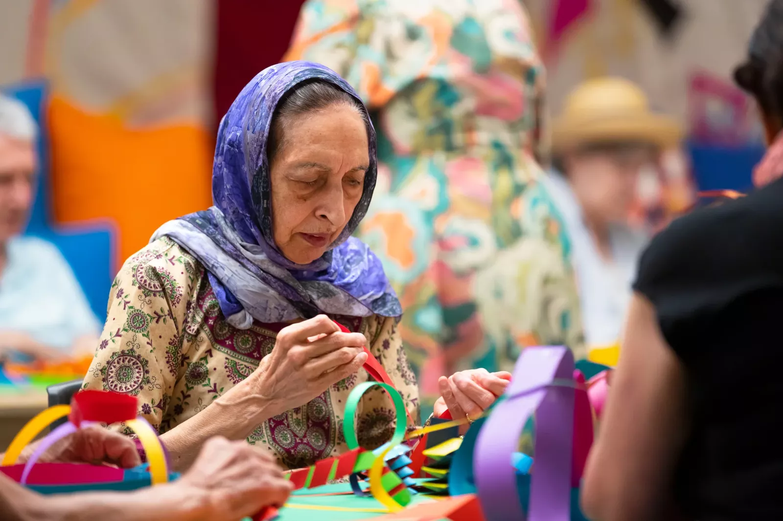 A woman wearing a head scarf is focussed on an artmaking task, assembling colourful strips of paper.