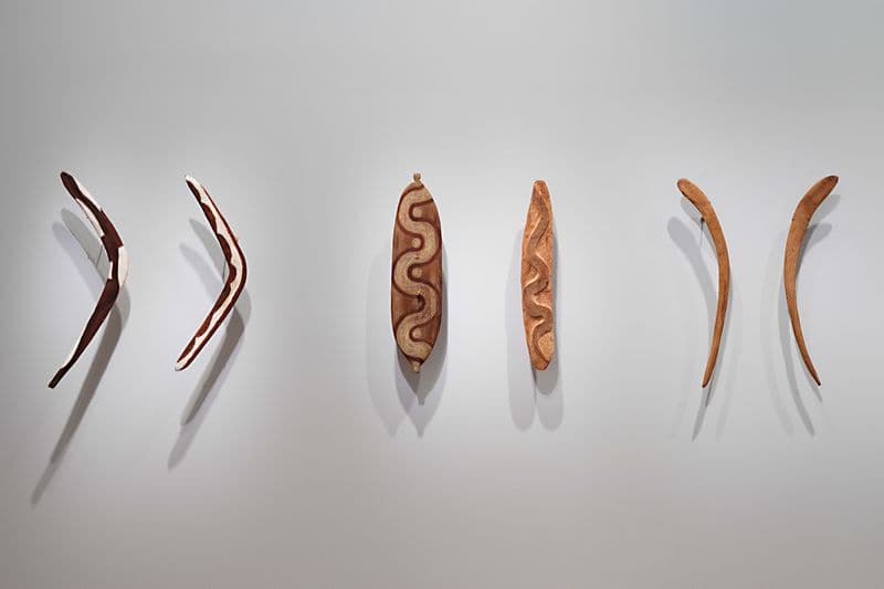 Four carved wooden boomerangs and two shields presented on a gallery wall