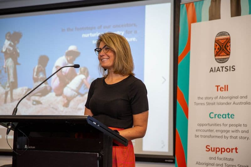 Female wearing glasses, with shoulder length blonde hair, wearing a black top and orange patterned skirt, standing at a lecturn