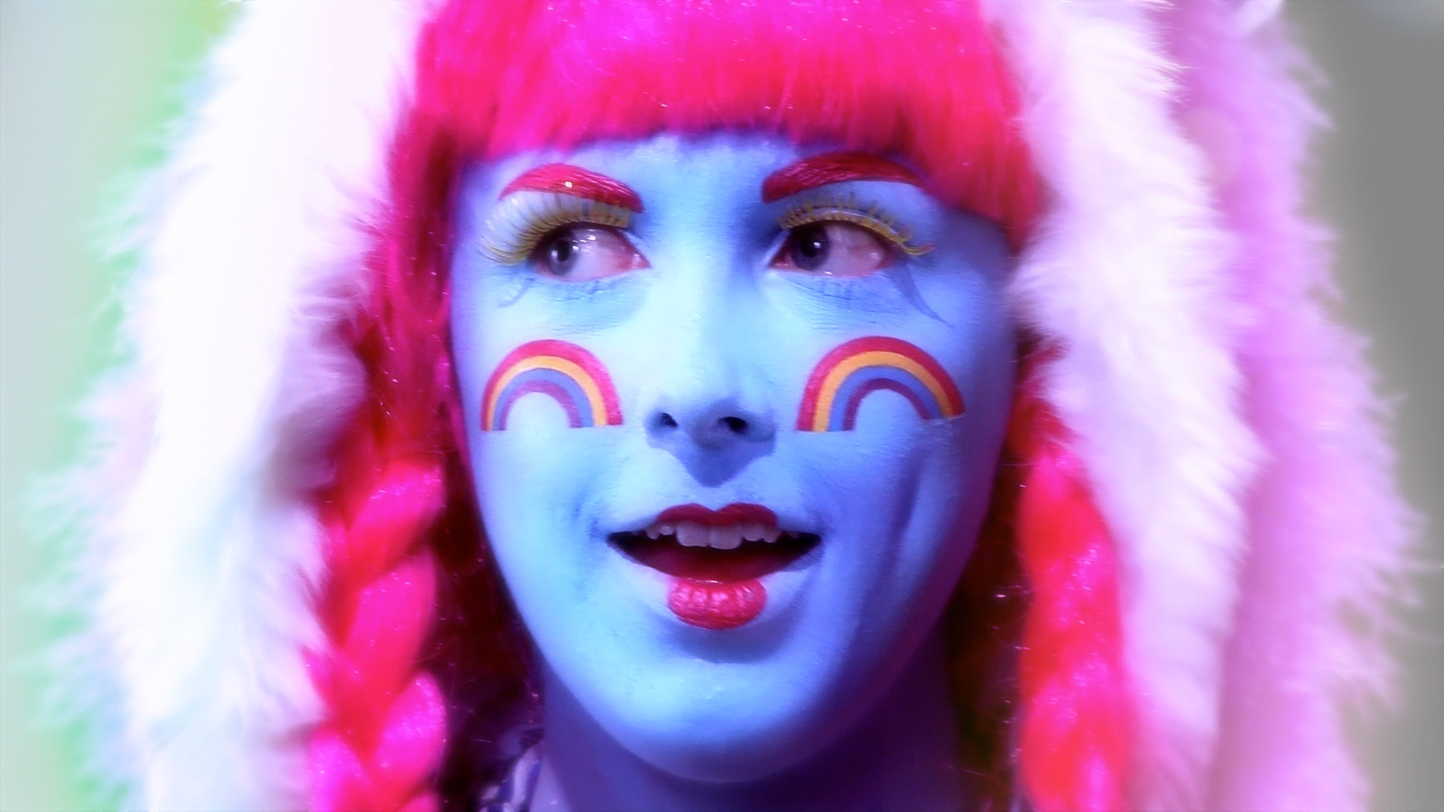 Close up of a woman's face with blue facepaint and rainbows on her cheeks with a bright pink wig