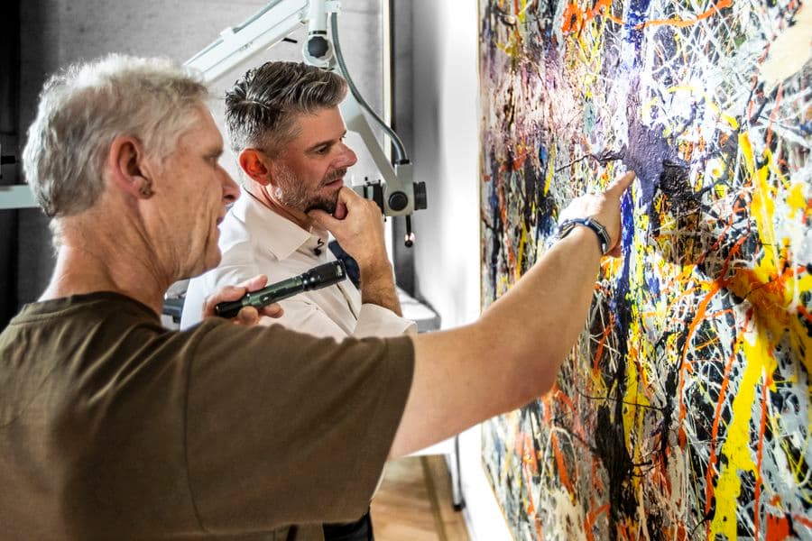 Photograph of conservator and director gesturing to painting under restoration