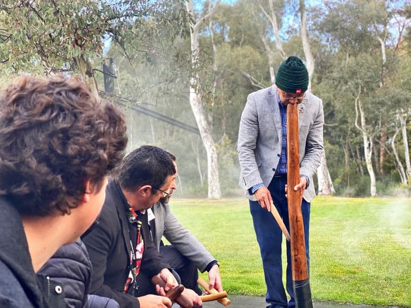 A man is standing up and playing the Didjeridu outside. Three other men can be seen seated next to him, watching and playing other instruments. There is smoke surrounding them.