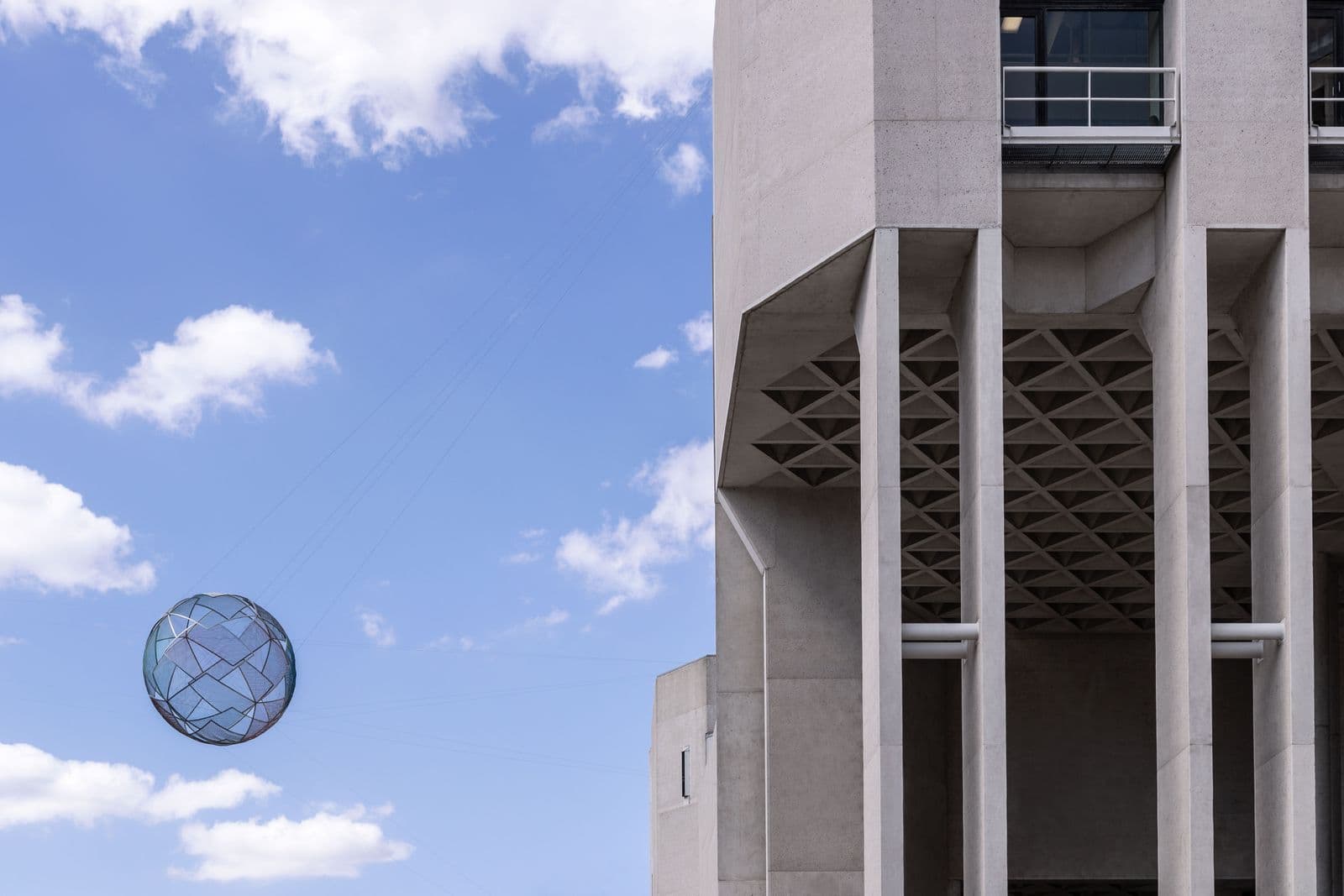 the edge of a large concrete brutalist building against a blue sky with a giant spherical sculpture suspended in the air