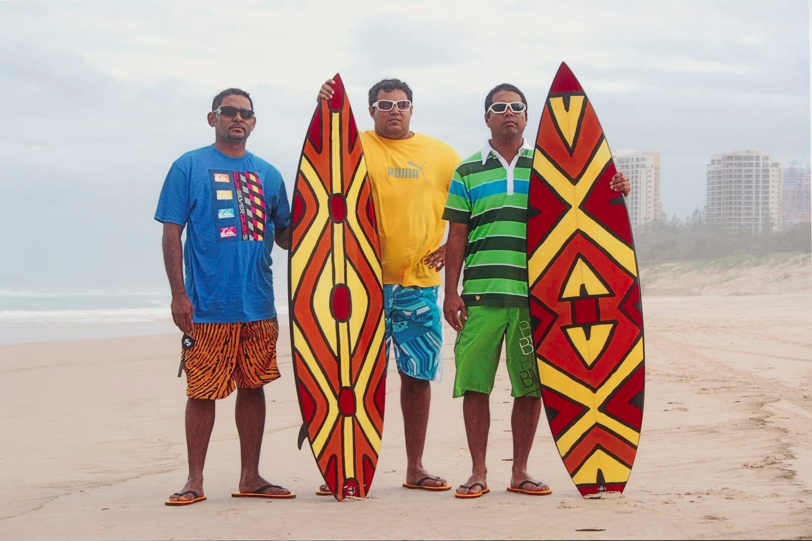 Three men pose on a beach with two coloured surfboards