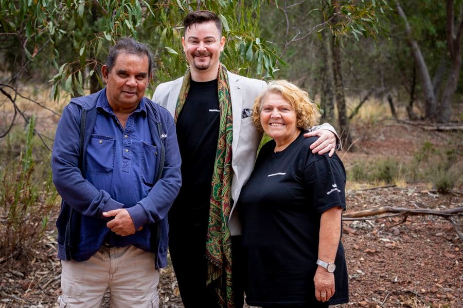 Photo of two men and a woman are standing outside beneath a gum tree. The man in the middle has his arms around the others, while they look at the camera smiling.