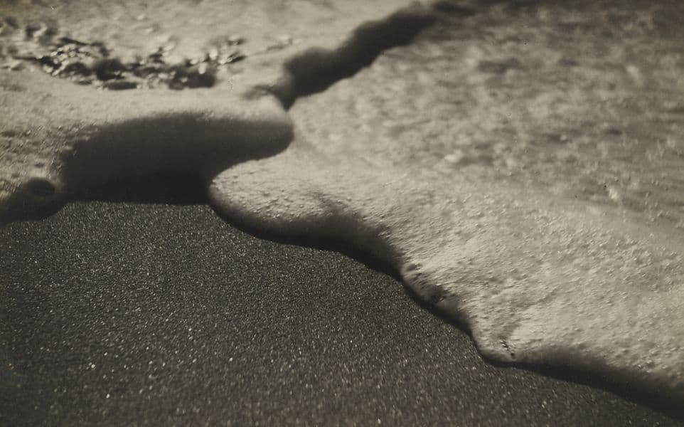 A black and white photo of the water's edge on a beach.