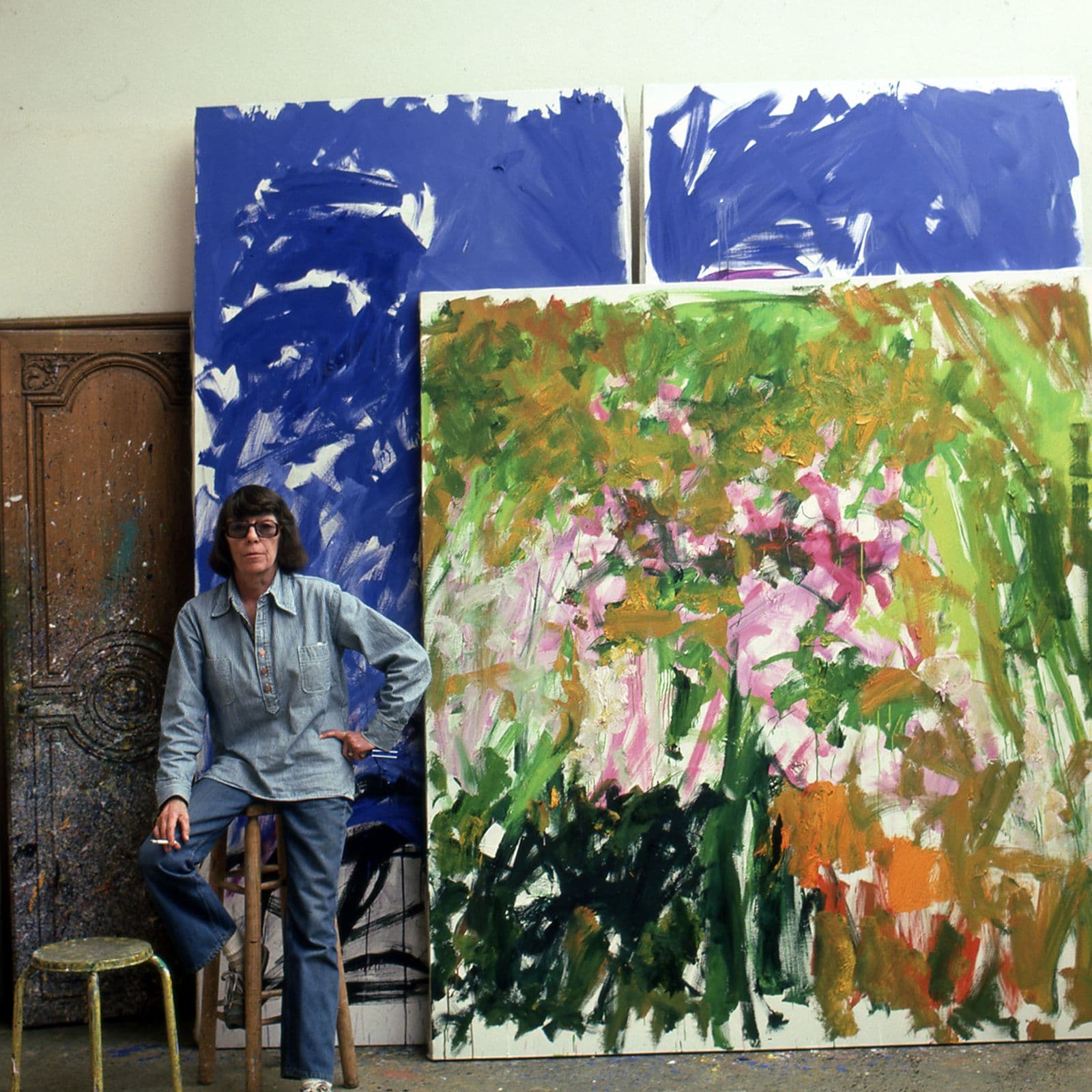 Joan Mitchell sits on a stool in front of two large painted canvasses