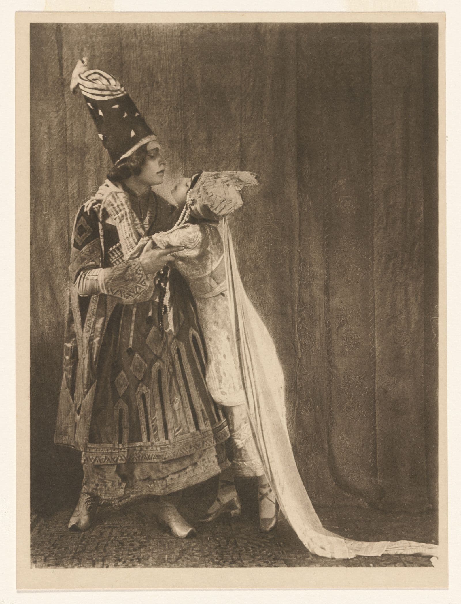 Photograph of two ballet dancers, a man and a woman, in a dramatic embrace. Both dancers are in elaborate costume.