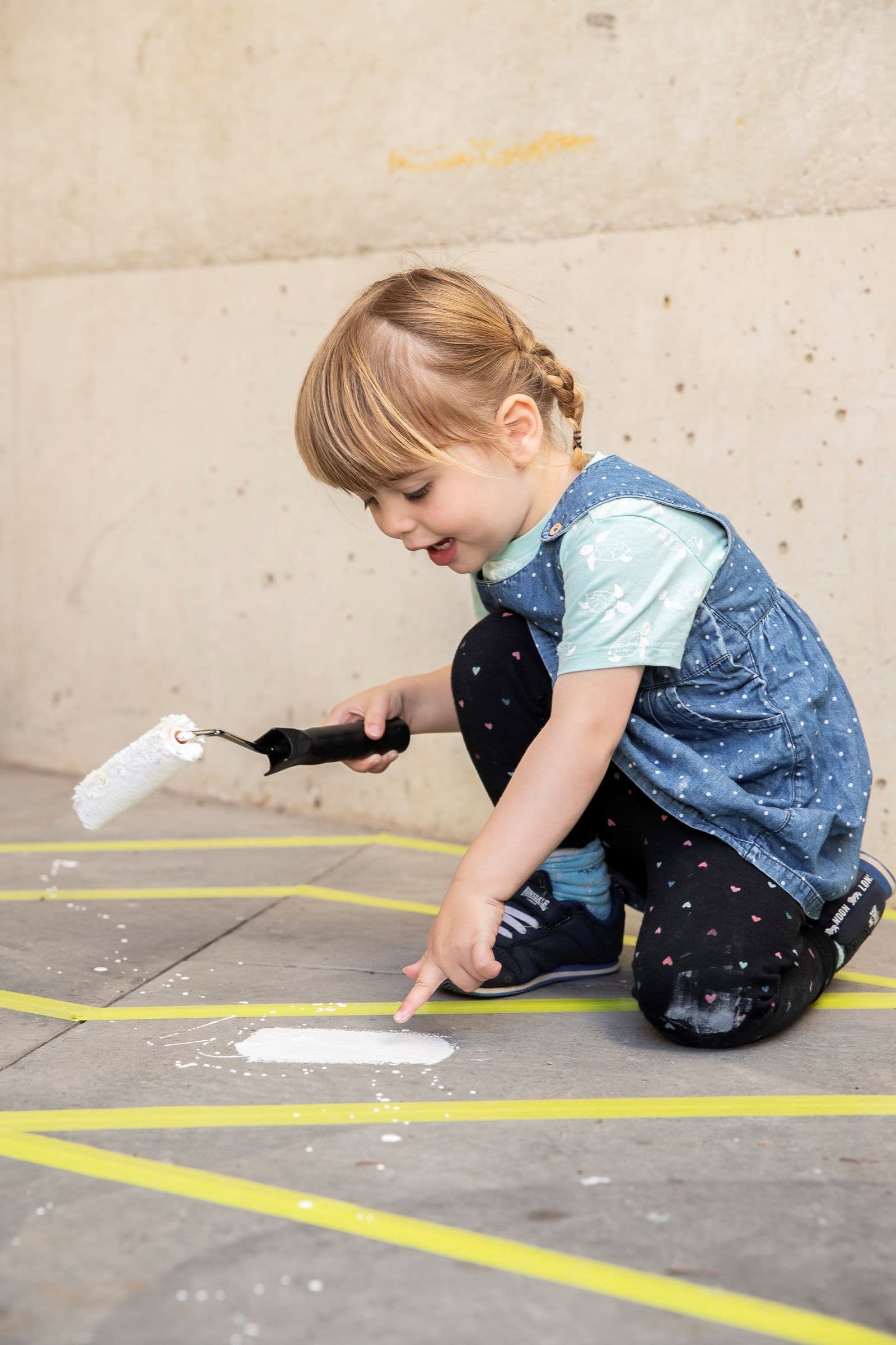 A young child is crouched down touching some paint they have put on the floor using a paint roller they are holding