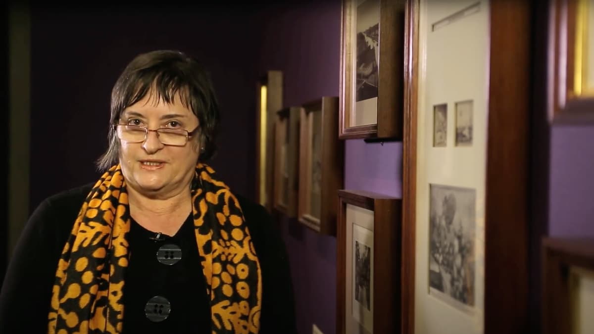 A video still of a woman speaking to the camera, standing in a gallery space