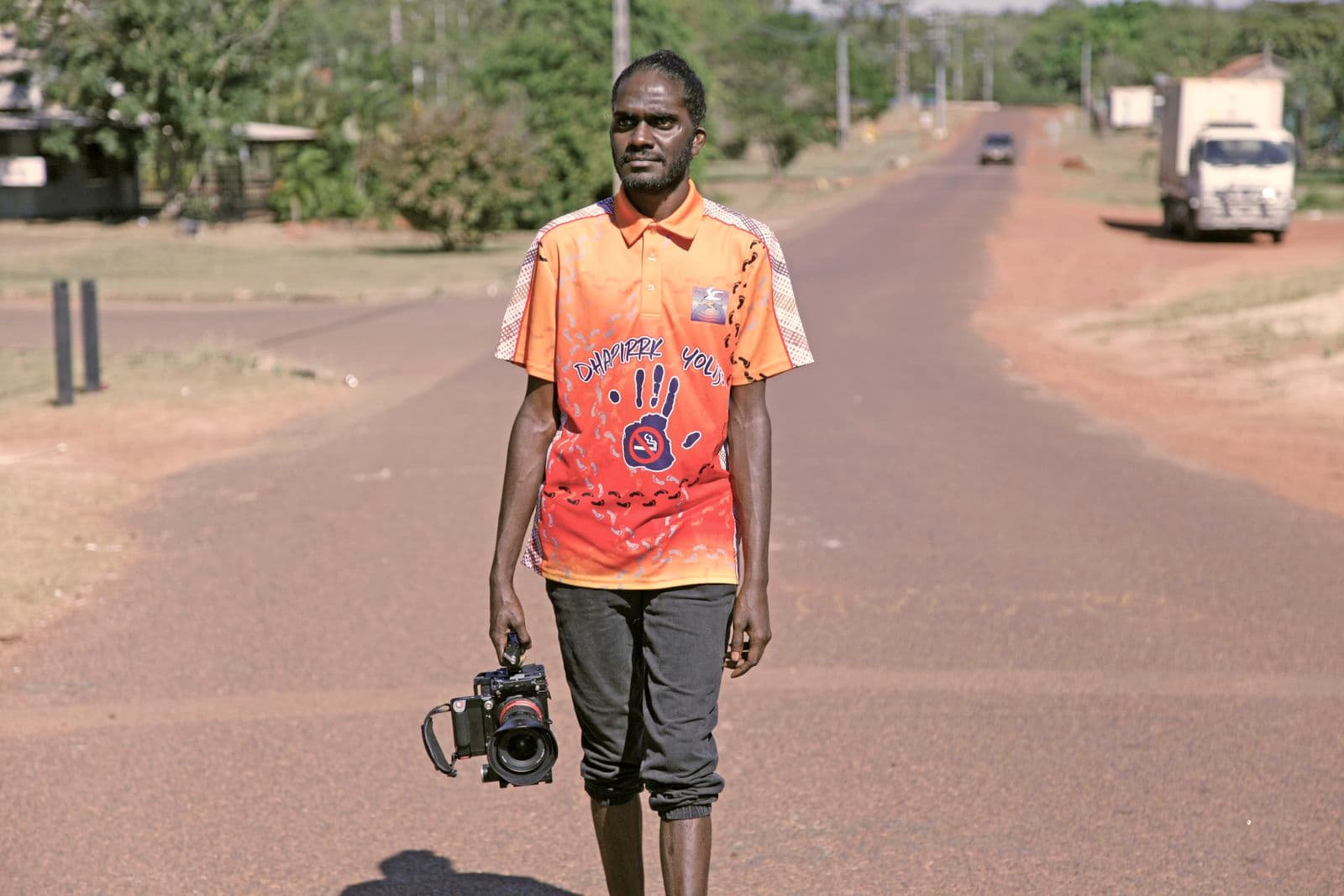 Artist Gutiŋarra Yunupiŋu carrying a camera and wearing a bright orange t-shirt walking walking through his hometown of Yirrkala in Northeast Arnhem Land