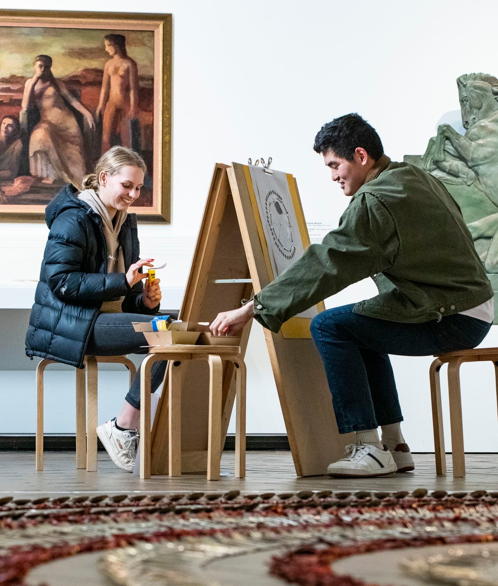 Two young people sit either side of an easel in a gallery space