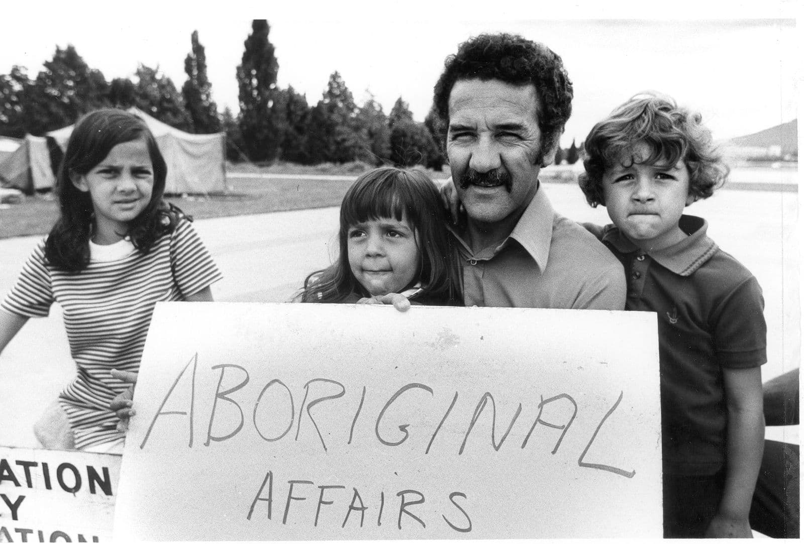 Photograph of three children with their father holding a sign stating "Aboriginal Affairs"