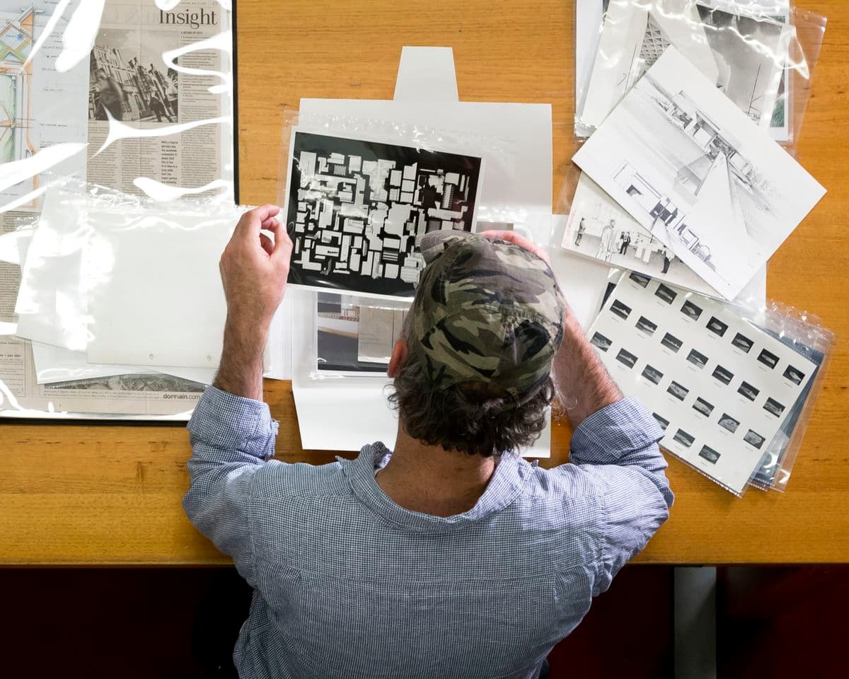 Photograph of a a white male wearing an army print hat rifles through building archives. Image is taken from behind.