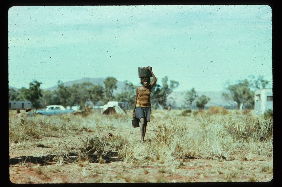Photo of a woman walking while carrying a container above her head and one in her hand in an outstation in the Northern Territory