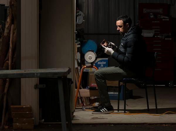 Lit by a single light, James Tylor sits in his darkened studio holding a daguerreotype photographic plate with gloved hands. He wears black and white Adidas trainers, olive chinos and a Kathmandu winter coat. He has dark short hair.