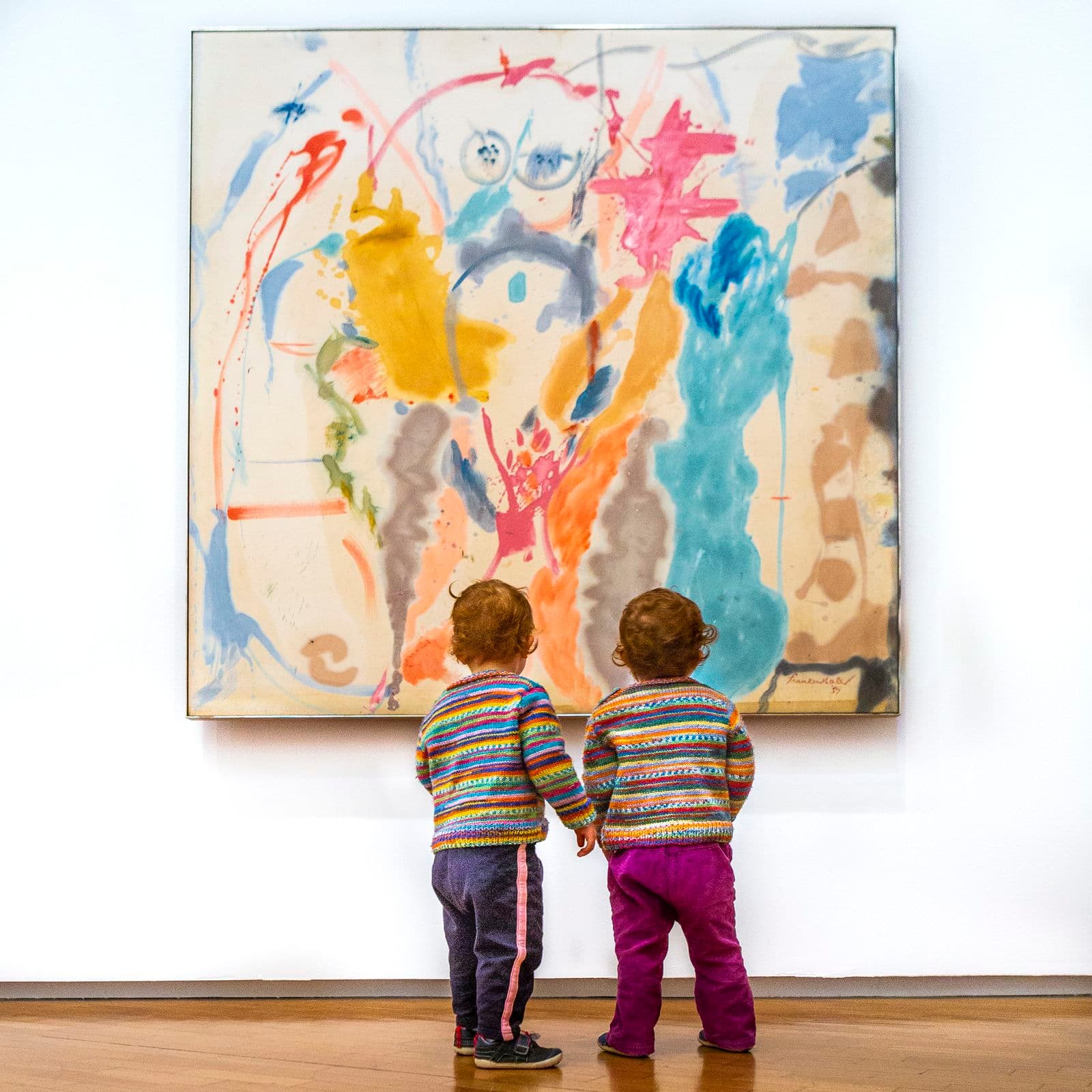 Two small children stand in front of a large painting